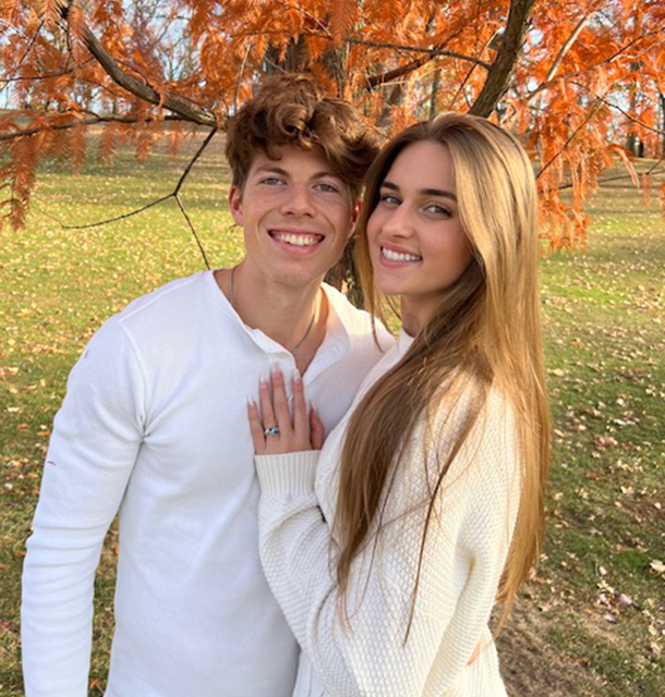 Two adults stand in a park in front of trees with colored foliage for fall